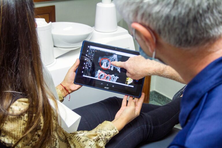 Patient sits in a dental chair while a dentist reviews a tooth X-ray beside treatment instruments.